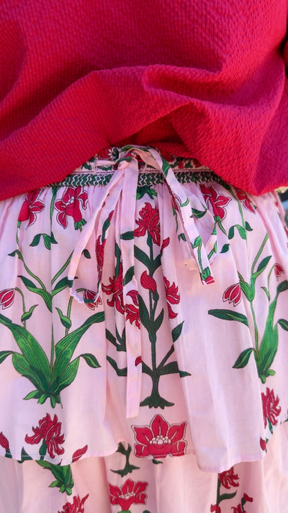 Rose d’Iris Ruffle Cascade Maxi Skirt, pink floral fabric with green and red patterns, tied at the waist with a matching ribbon; available in sizes XS–XL.