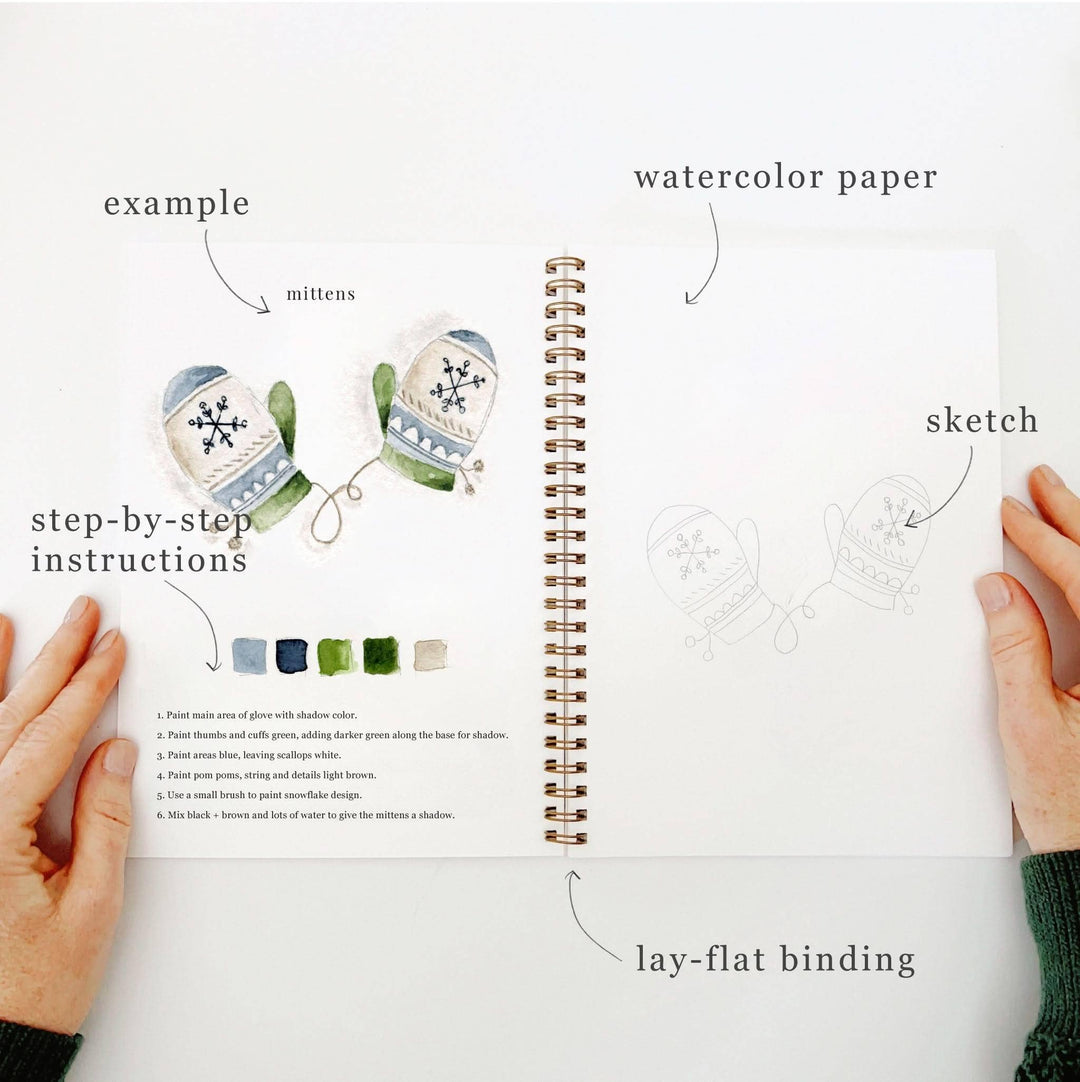 Winter watercolor workbook open to a page with mitten illustrations, featuring a painted example, a pencil sketch, and step-by-step instructions.