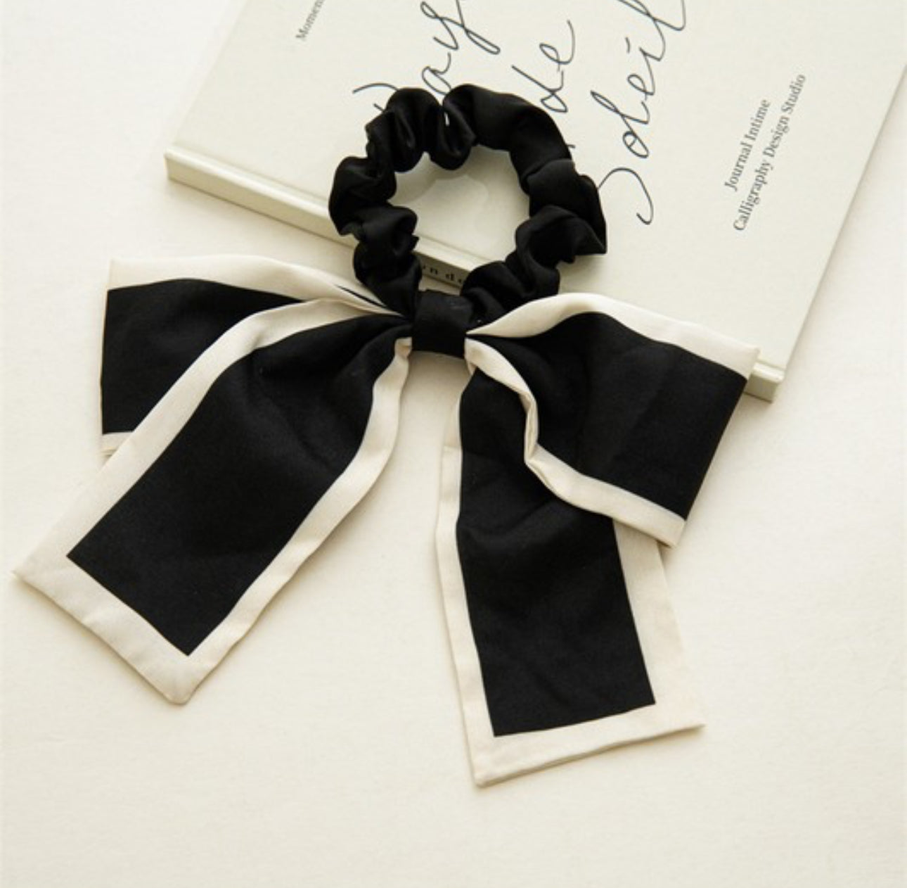 The Favola Bow Hair Tie is a stylish accessory in black and white. This desirable hair tie features elegant, long ribbon tails, adding a playful yet sophisticated touch to your hairstyles. Available in one size fits all.