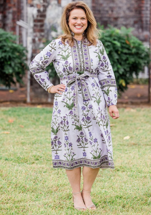The Iris Lavende The Caroline Maxi is a long-sleeved, knee-length cotton poplin dress with a purple floral pattern, hand block printed details, and a tie at the waist. Available in sizes XS-XL.