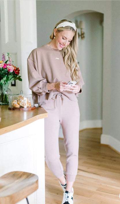 A woman in a pink Adidas Originals Millie LUX Sweatshirt and white sneakers smiles while looking at her phone, standing near a kitchen counter with flowers and cookies.