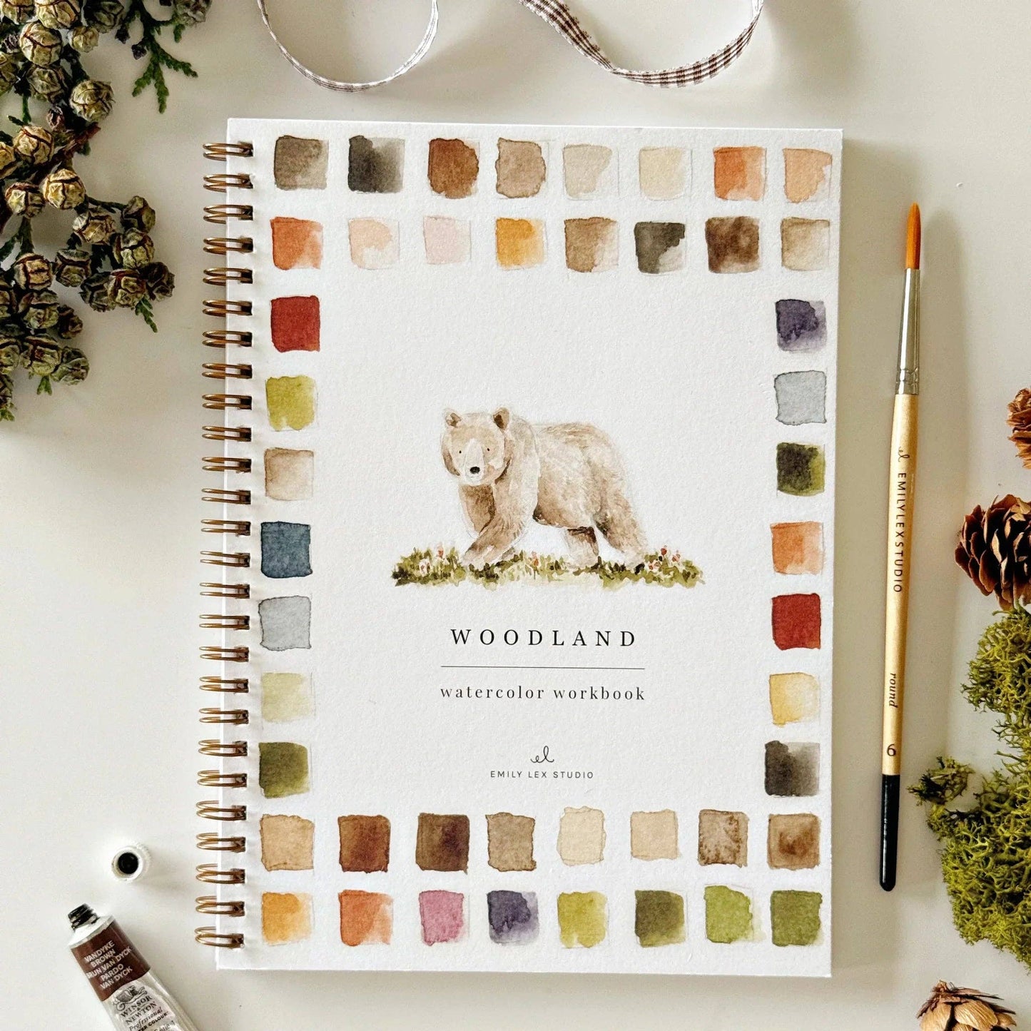The Woodland watercolor workbook is a spiral-bound painting guide with a charming bear illustration on the cover, framed by vibrant paint swatches. Available in multiple sizes. Includes step-by-step instructions and woodland illustrations.