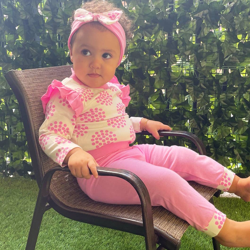 A toddler girl with a pink headband sits on a patio chair against a green artificial hedge background, wearing a Layette Girls Baby clothes Shower Gift Pink Polka Dot Easter: 3-6 Mo outfit.