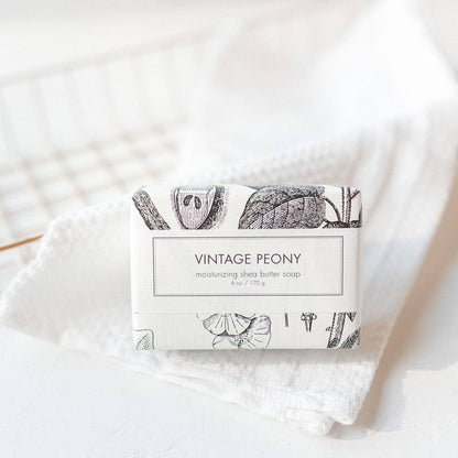 Formulary 55's Vintage Peony Soap is a luxurious bath bar infused with rich Shea butter. Presented in an artistic box adorned with botanical illustrations, it accentuates any bathroom decor. The soap exudes a soft, floral scent appealing to every sense. It lies beautifully on a light textured surface under soft sunlight which enhances its physical appeal. Available only in one standard size for maximum indulgence.