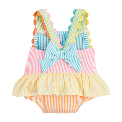 A vibrant baby dress with pink, blue, yellow, and orange stripes, featuring scalloped crossed straps, a large back bow, and a ruffled skirt. Available sizes: 3M, 6M, 9M, 12M.