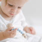A toddler wearing a white shirt closely examines a pink and blue Pacifier Clip. The child is focused, and the background is bright and softly blurred. The beads are made from food-grade silicone.