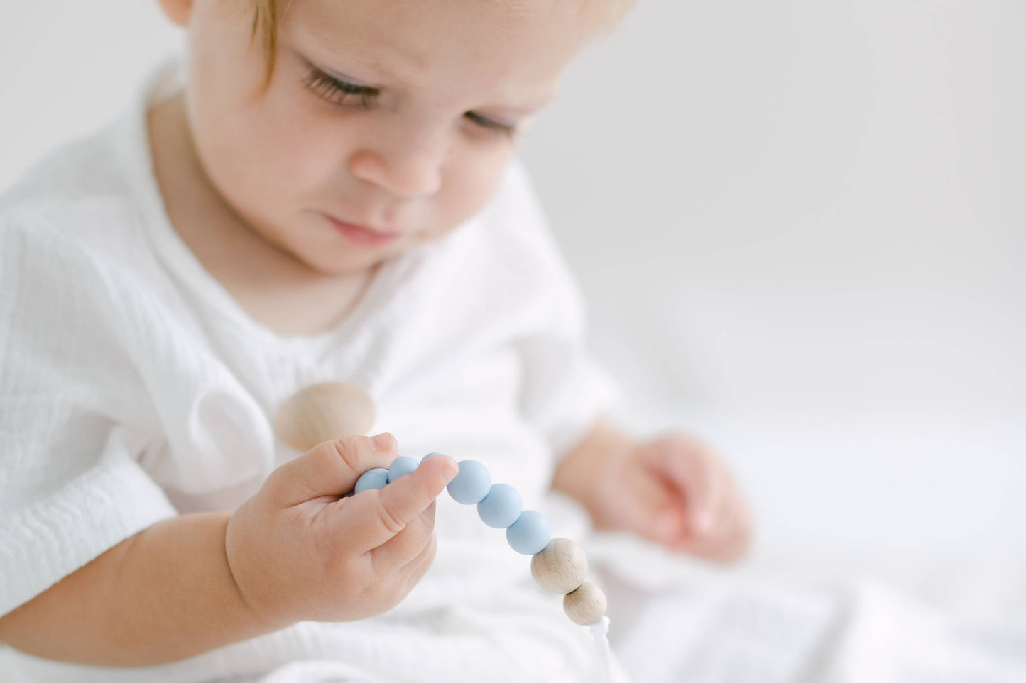 A toddler wearing a white shirt closely examines a pink and blue Pacifier Clip. The child is focused, and the background is bright and softly blurred. The beads are made from food-grade silicone.