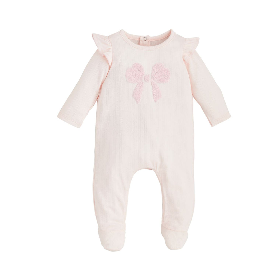 The Ruffle Bow Baby Sleeper is a light pink, soft cotton rib onesie with long sleeves and footed bottoms. It features a textured bow on the front and ruffle details on the shoulders. Available sizes: newborn, 0-3 months, 3-6 months.