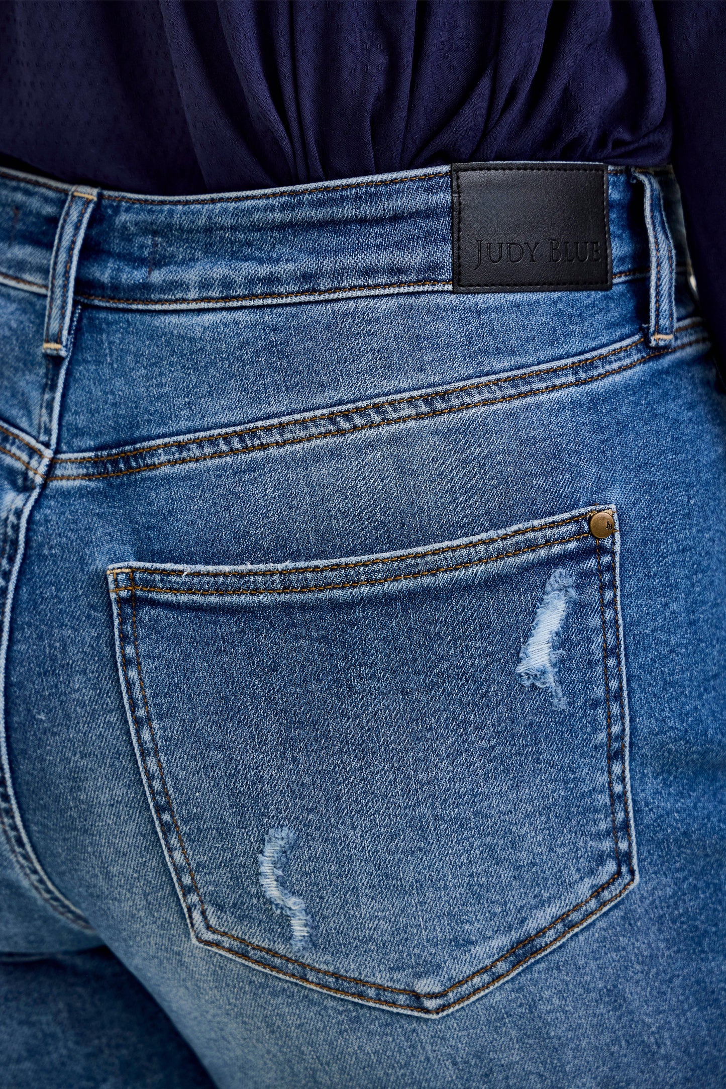 Close-up of the back pocket and waistband of the Billie HW R.M. Boyfriend Classic Stone Blue Jeans in medium stone blue wash with distressed details and a black “Judy Blue” patch. High waisted boyfriend style, rigid magic construction for comfort and style. Available in sizes 0-24.