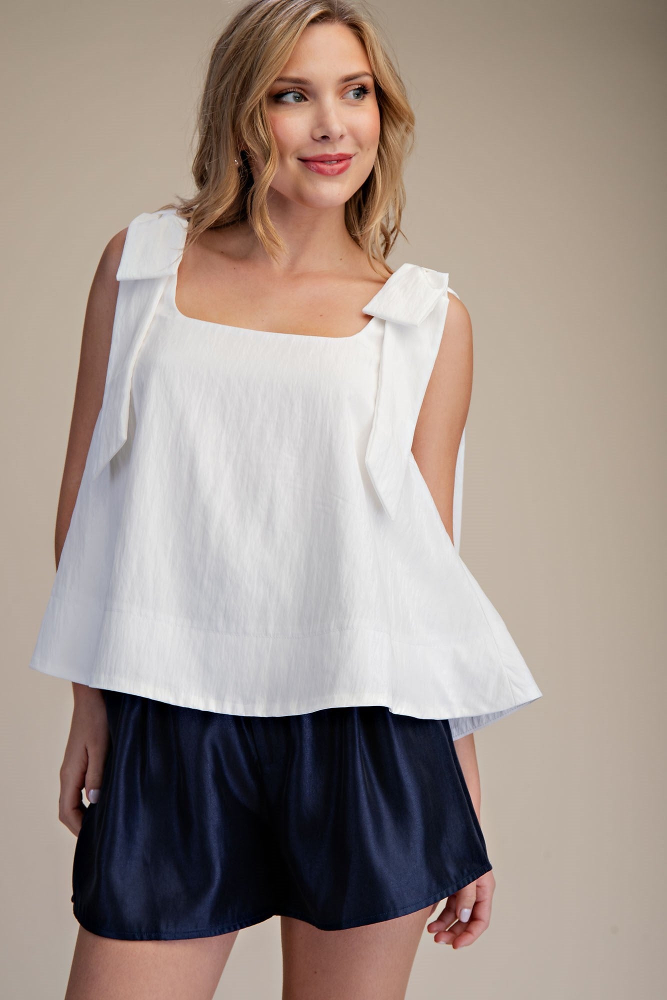 Sleeveless white scoop neck top with bow shoulder detail. Available in XS-XL. Navy blue shorts not included.
