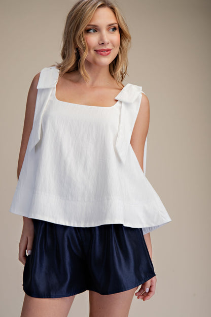 Sleeveless white scoop neck top with bow shoulder detail. Available in XS-XL. Navy blue shorts not included.