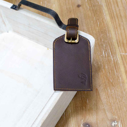 The Leather Embossed Luggage Tag features genuine brown leather, a subtle embossed logo, and a brass buckle. Available size: One Size.