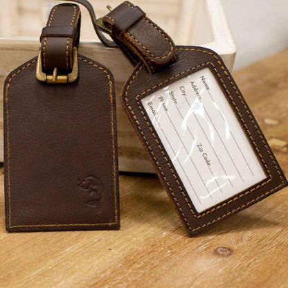 Leather Embossed Luggage Tags come in two designs: one with a stitched pattern and the other with a clear plastic window for personal info. Both include brown stitching and buckle straps. Sizes: Not specified.