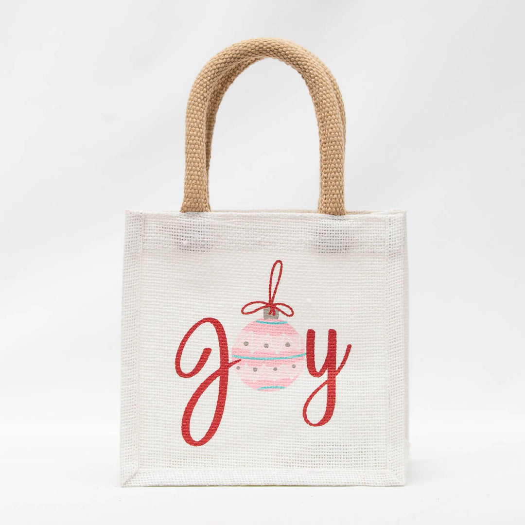 Holiday Petite Gift Tote, a small white reusable mini gift tote with woven brown handles featuring a red embroidered “joy” with a pink and blue striped ornament as the letter “o.”.