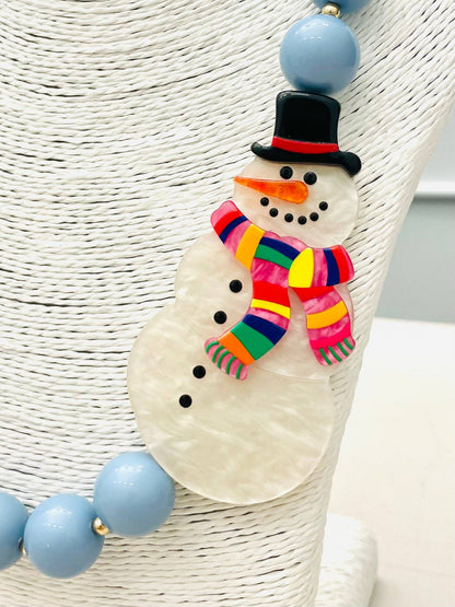 Beaded Snowman Necklace (blue/white) BNK1216 features a snowman pendant with a black hat, orange nose, striped scarf, and black buttons on large light blue beads. Available sizes: S, M, L.