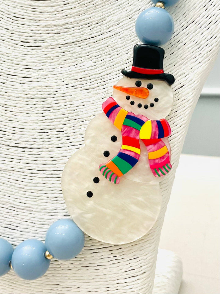 Beaded Snowman Necklace (blue/white) BNK1216 features a snowman pendant with a black hat, orange nose, striped scarf, and black buttons on large light blue beads. Available sizes: S, M, L.