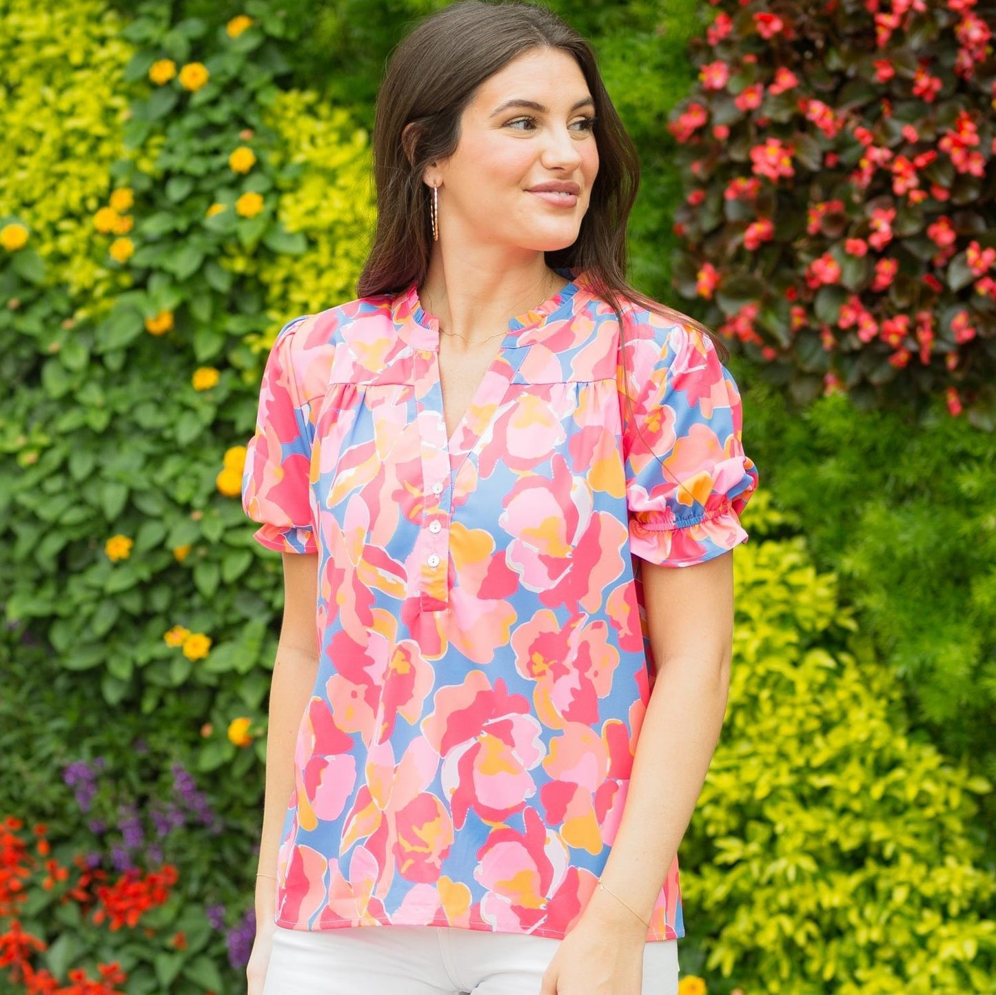 A woman in a colorful floral Sutton Top - Plus smiles and looks to her side, standing before a vibrant garden with multicolored flowers, illustrating the sizing.