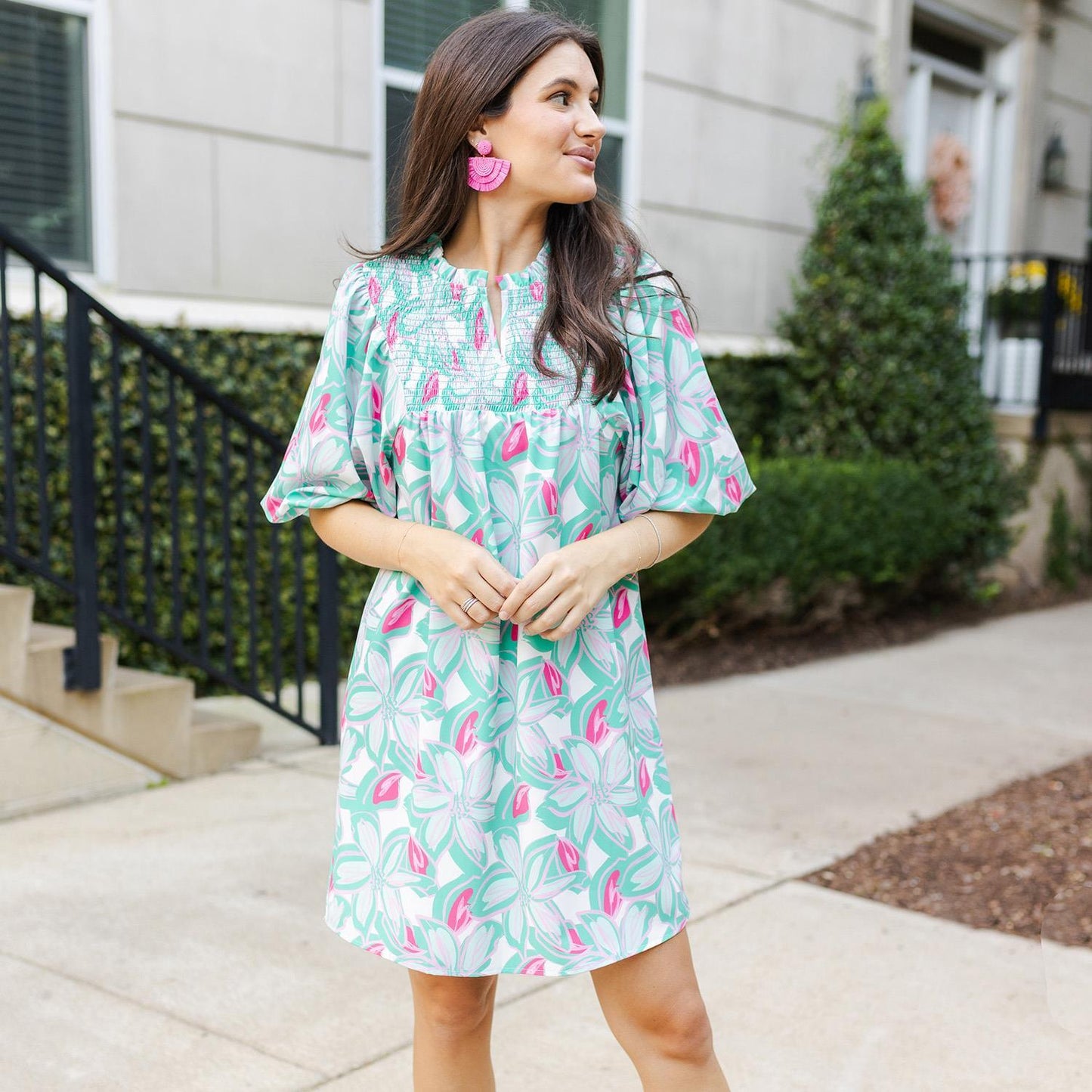 The Amelia Paradise View Mint Dress is available in small, medium and large sizes. It features a green and pink floral pattern throughout the design, perfect for any summer occasion. It is complemented with large pink earrings to complete the look.