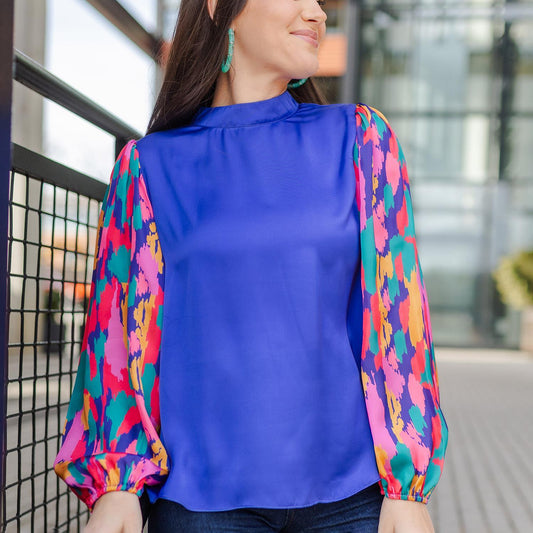 Melanie Stroke of Luck Purple Top featuring colorful, abstract-patterned long sleeves in shades of pink, yellow, purple, and green. Sizes available: XS-XL.