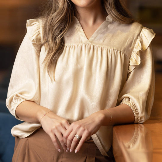 The Parker Top is a cream-colored festive blouse with ruffled sleeves and an elegant shimmer. Available in sizes XS, S, M, L, and XL.