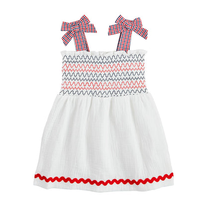 The White Smocked Gauze Toddler Dress is a charming cotton outfit with smocked zigzag patterns in red and navy, gingham bow straps in red and blue checks, and a red wavy hem trim. Available sizes: 12M, 18M, 24M, 2T, 3T.