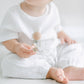 A toddler in white clothing sits on a pale surface, holding a pink and blue Pacifier Clip, with a focus on the child's bare feet and the clip in hand.