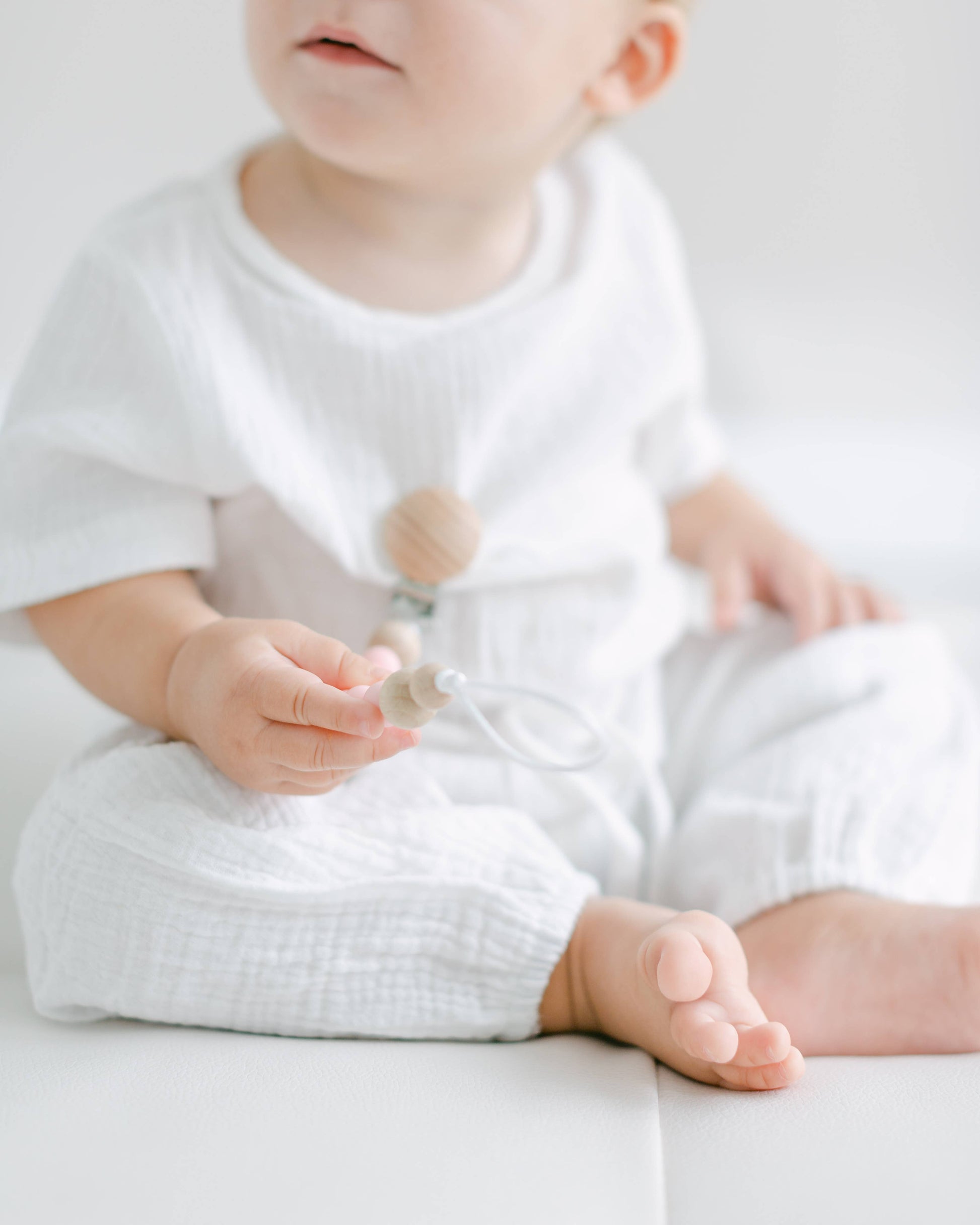 A toddler in white clothing sits on a pale surface, holding a pink and blue Pacifier Clip, with a focus on the child's bare feet and the clip in hand.