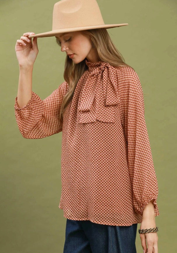 The Dottie Tie Top is a patterned blouse featuring a bow at the neck, available in sizes XS-XL.