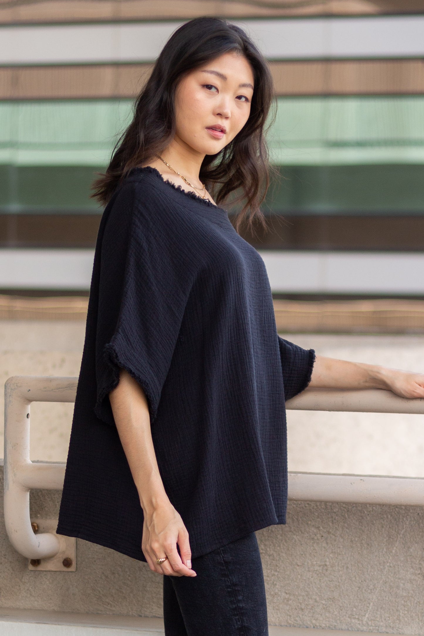 A woman in a Gauze Raw Edge Poncho Top and jeans posing with one hand on a railing, looking away from the camera, with a blurred building background.