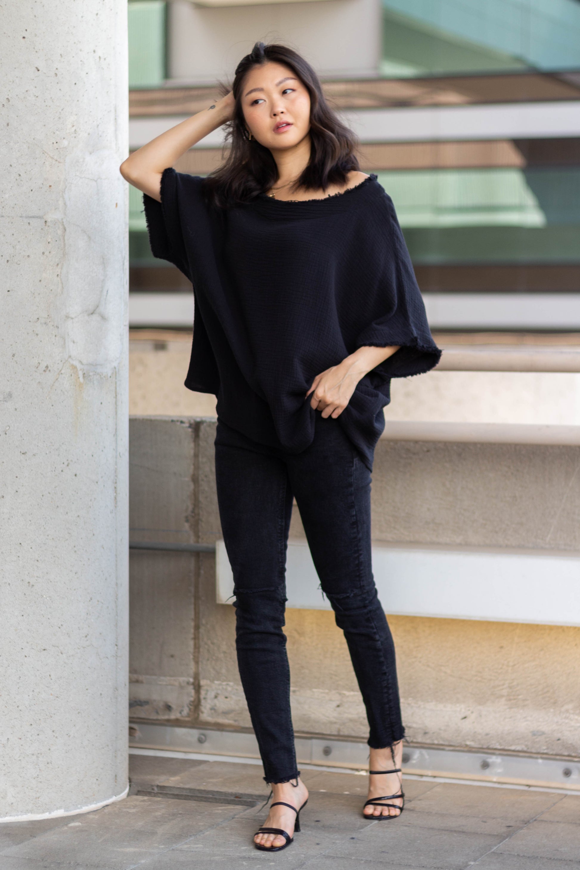 A woman in a stylish black Gauze Raw Edge Poncho Top outfit, standing in an urban setting, poses with one hand on her head and a thoughtful expression.