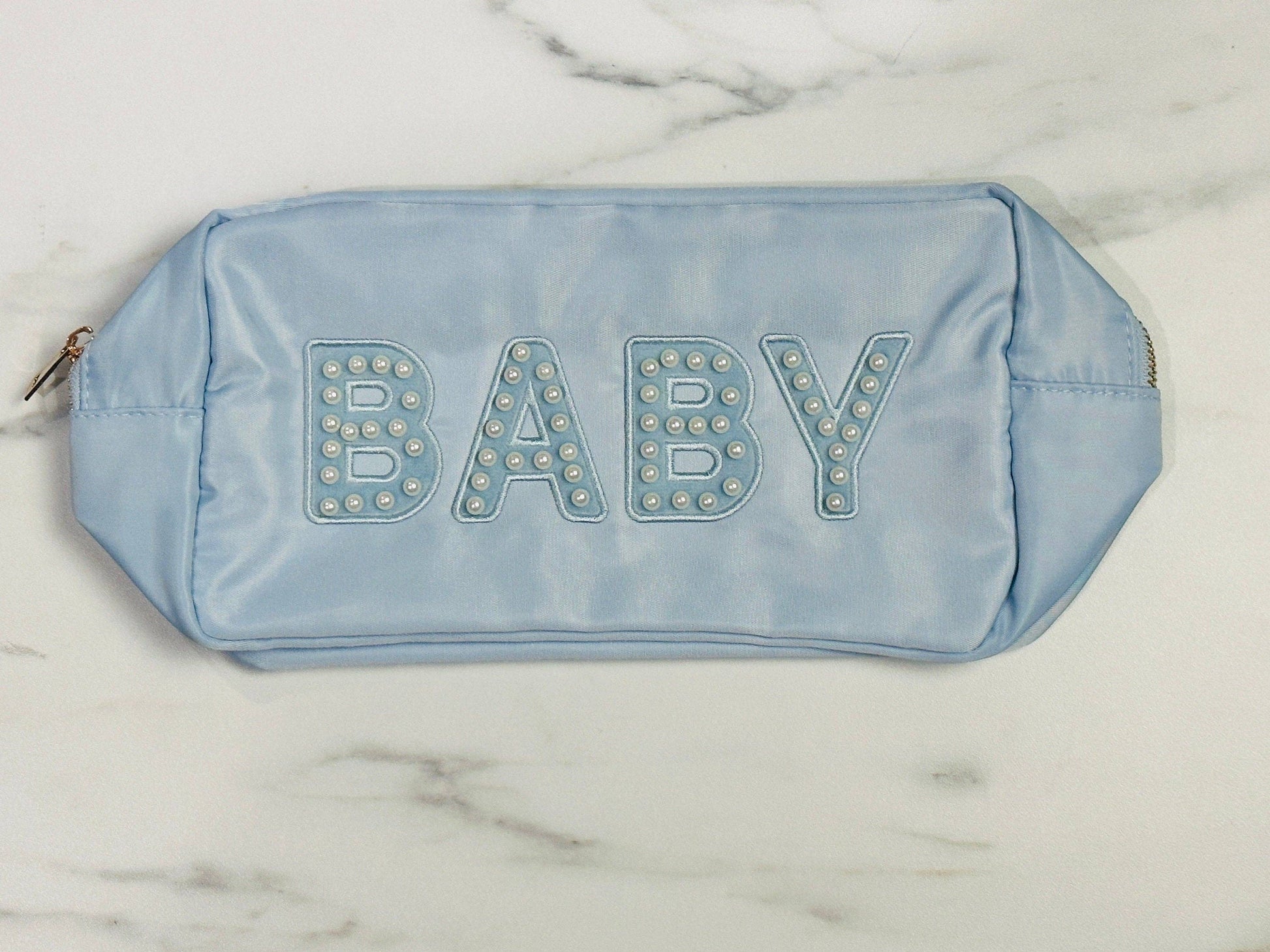 The Baby Pearl Varsity Letter Pouch is a light blue rectangular nylon bag with "BABY" embroidered in large letters on the front, adorned with small pearl-like beads. It features gold zippers on both ends. Sizes available: Small, Medium, Large.