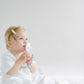 A toddler with light hair in a white outfit sitting on a white backdrop, looking upwards thoughtfully while holding a Pink and Blue Pacifier Clip.