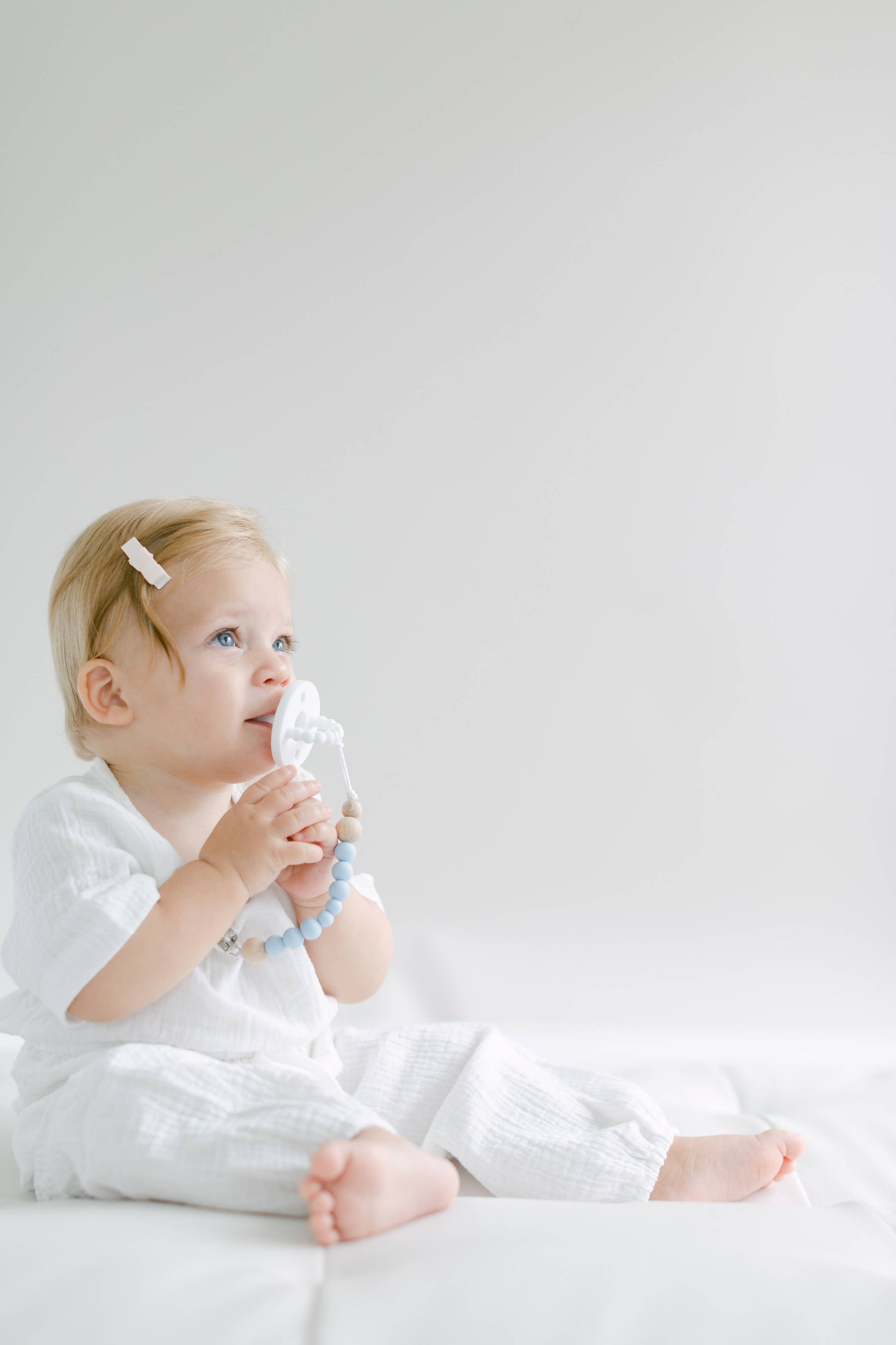 A toddler with light hair in a white outfit sitting on a white backdrop, looking upwards thoughtfully while holding a Pink and Blue Pacifier Clip.