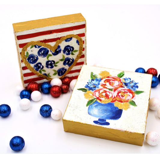 The product consists of two 4x4 handpainted wood block signs. The first sign features a vibrant square floral motif while the second sports a unique heart-shaped design. Both are adorned with dotted red and blue decorative balls set against a pristine white backdrop. Ideal for summer celebrations, these pieces bring an artful blend of color and charm to any setting.