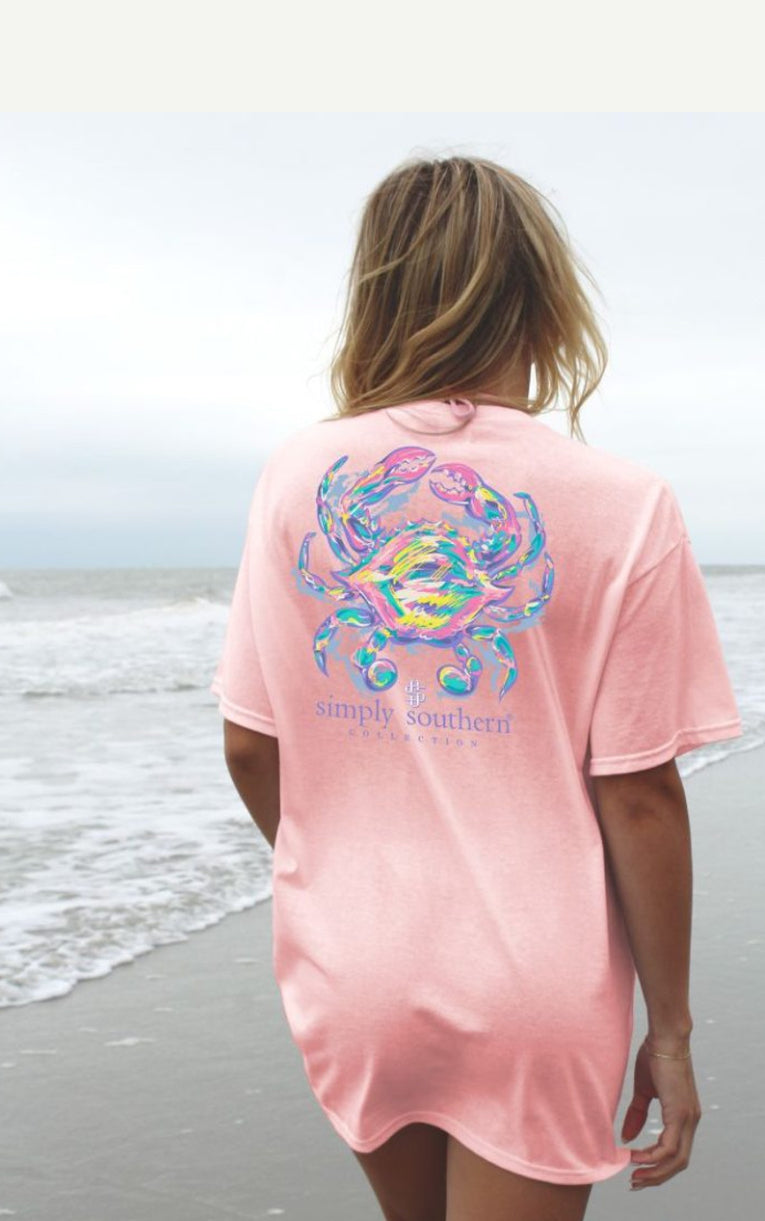 A woman stands on a beach facing away from the camera, wearing a pink short sleeve shirt with a colorful crab Lotus design and the text "Simply Southern" printed on the back.