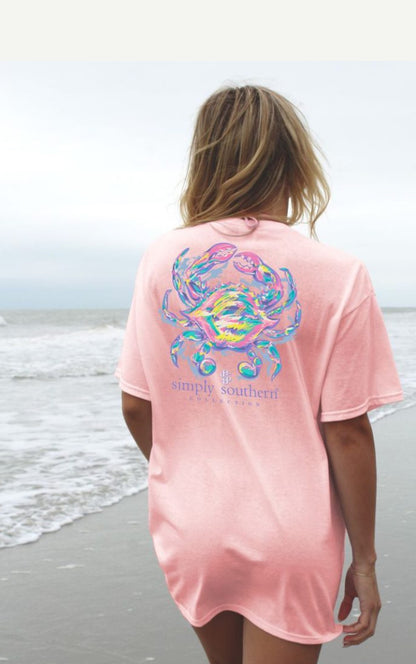 A woman stands on a beach facing away from the camera, wearing a pink short sleeve shirt with a colorful crab Lotus design and the text "Simply Southern" printed on the back.