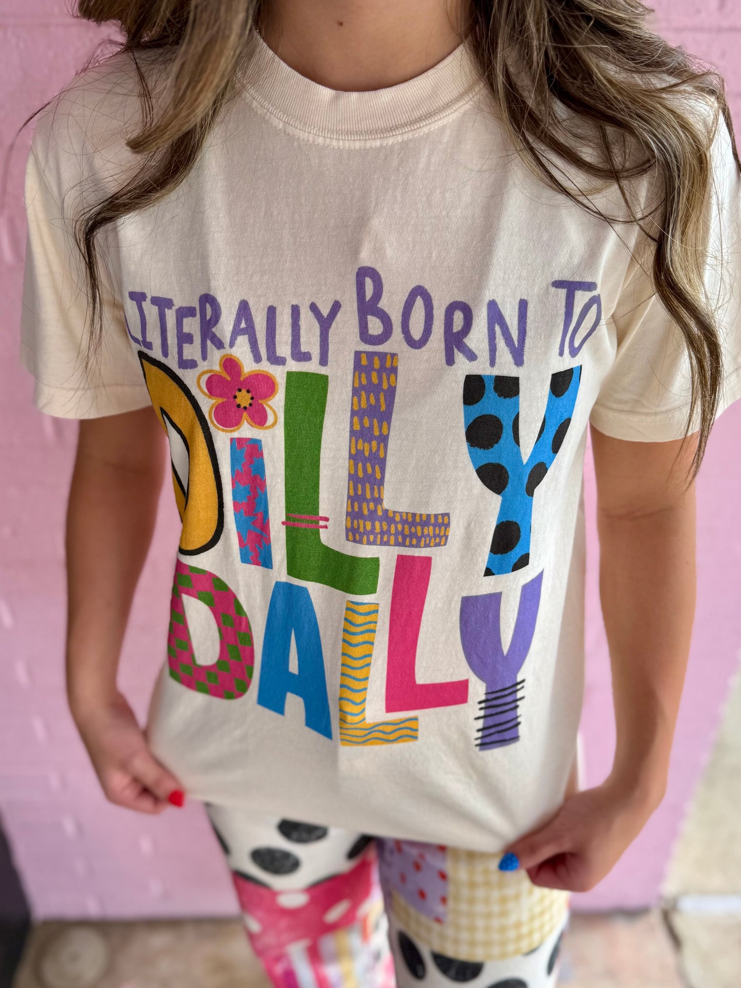 Cream garment-dyed cotton tee with "Literally Born to Dilly Dally" graphic. Sizes: Youth XS–XL, Adult S–3XL.