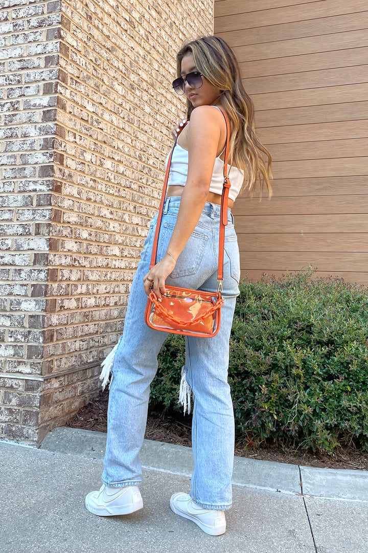 Jessica Clear Crossbody handbag: Transparent body with white trim, adjustable crossbody strap, top zipper closure. Available in one size: 8” W x 6” H x 2.5” D.