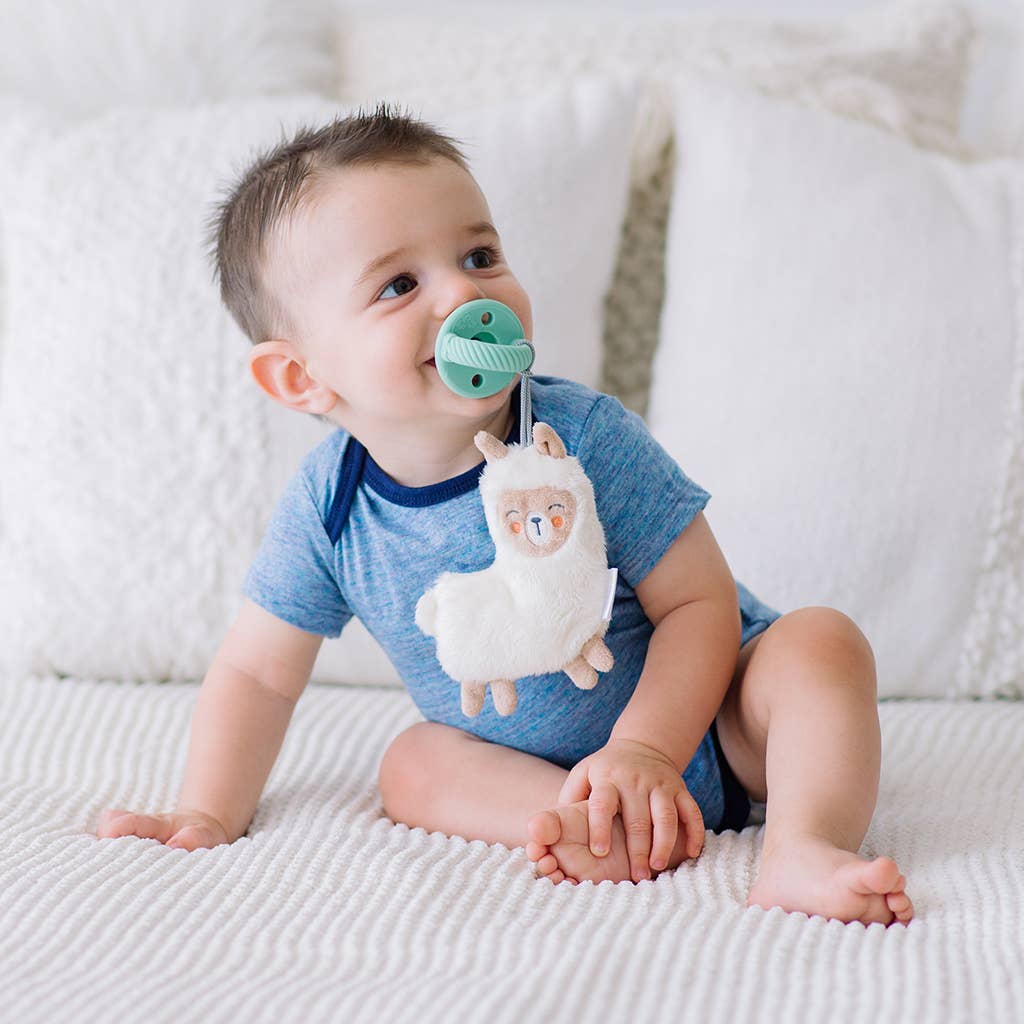 A baby sits on a white blanket, wearing a blue shirt and using a Sweetie Pal™ Plush & Pacifier: Dino, looking up with a curious expression. White pillows are in the background.