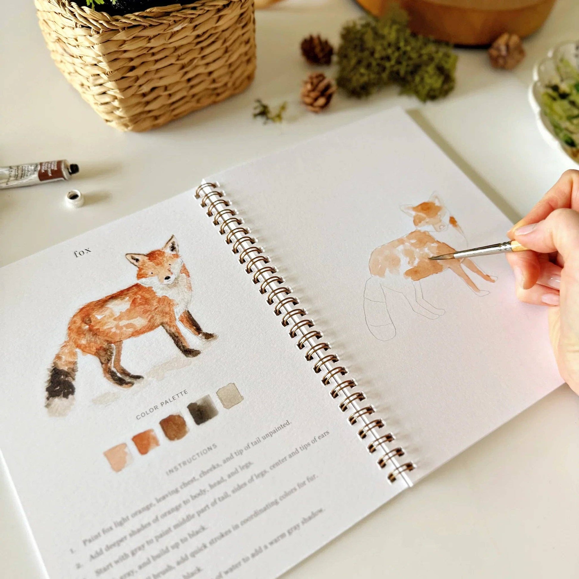 The Woodland Watercolor Workbook lies open, showing a fox painting tutorial for beginners. The right page displays a fox in progress, while the left has a completed fox and color palette. Available in multiple sizes.