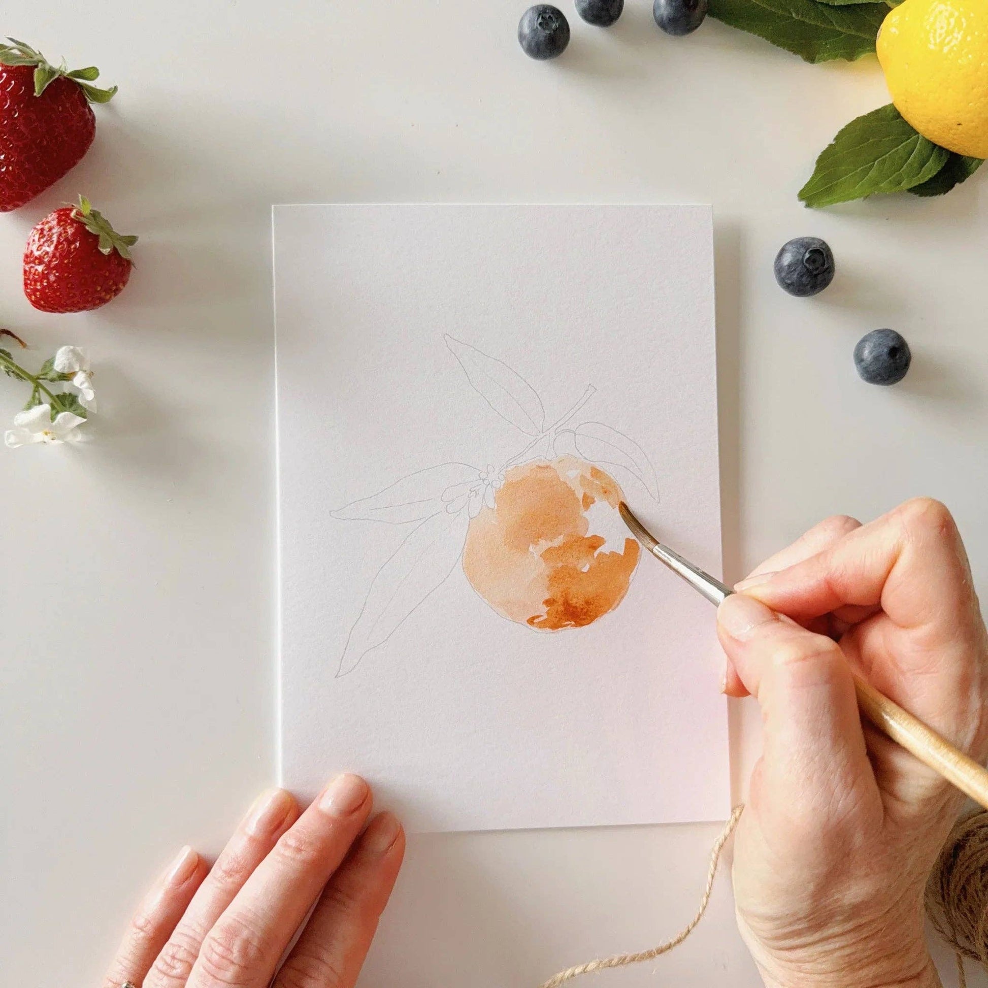A hand paints an orange fruit on a white card from the Fruit Paintable Watercolor Notecard Set, available in multiple sizes.