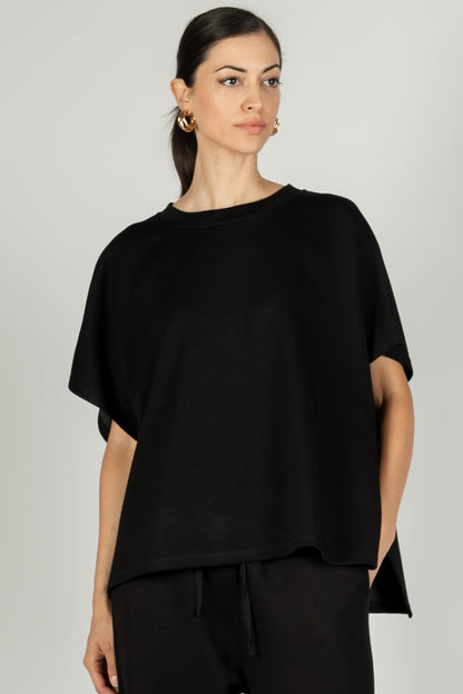 A woman with her hair pulled back wears a P. CILL Butter Modal Side Slit Cape Top paired with matching black pants, showcasing the sizing.