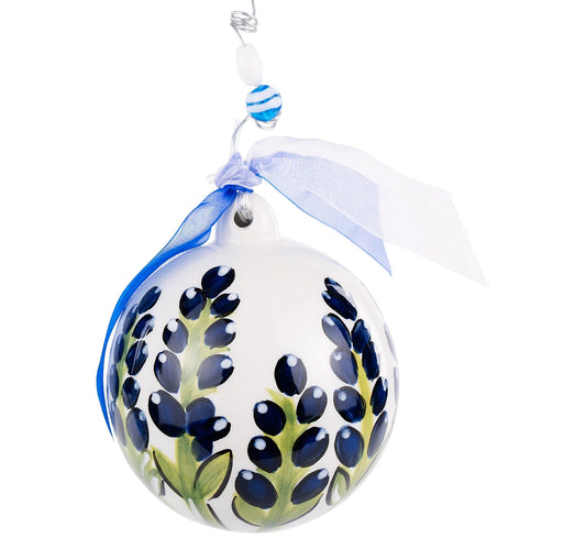 The Blue Bonnet Ornament is a spherical white Christmas ornament with hand-painted bluebonnet flowers and green leaves. It includes a decorative blue and white ribbon, a blue and white striped bead, and hangs from a metallic hook. Available in one size.