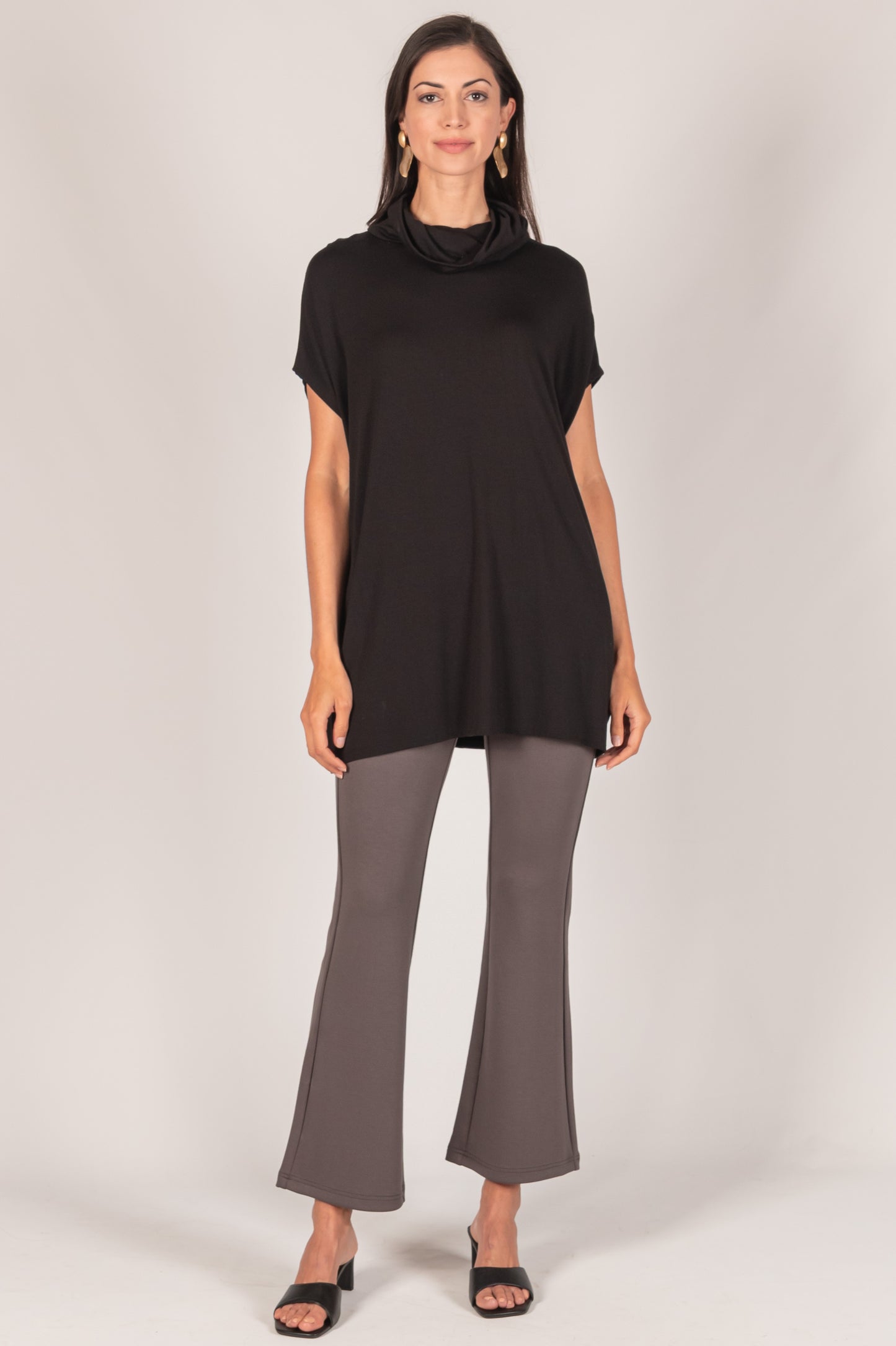 A woman wears a stylish outfit featuring a Super Rayon Jersey Cowl Neck Tunic with a draped neckline and gray flared pants, paired with black open-toe heels. Sizing options are available.