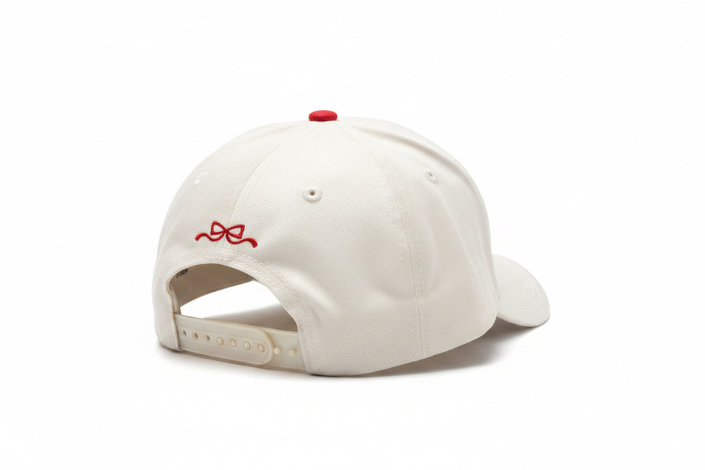 Cream-colored baseball cap with adjustable strap and small red bow embroidery above the back opening. Available in sizes S, M, and L.