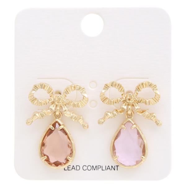 Gold-toned bow earrings featuring rhinestones and dangling teardrop gemstones in light brown and pale pink. Presented on a white backing card labeled "Lead Compliant." Size: One size fits all.