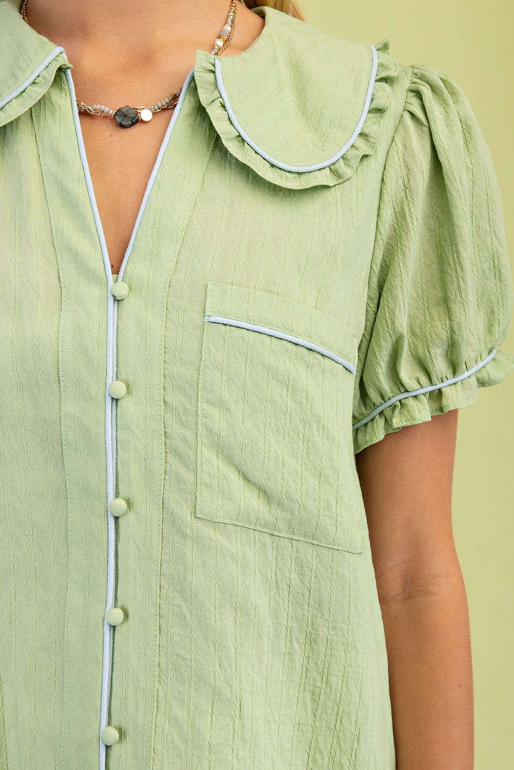 Light green blouse with a ruffle-trimmed Peter Pan collar, puff sleeves, light blue piping on front and pocket, small buttons. Sizes: XS, S, M, L, XL.