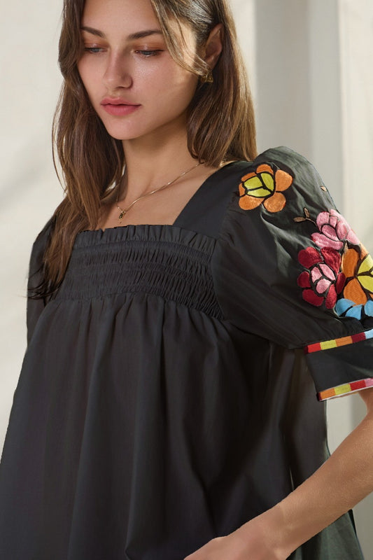 Daisy Sunset Embroidery Top: Loose black top with square neckline, gathered details, and short sleeves featuring colorful floral embroidery. Available in sizes XS–XL.