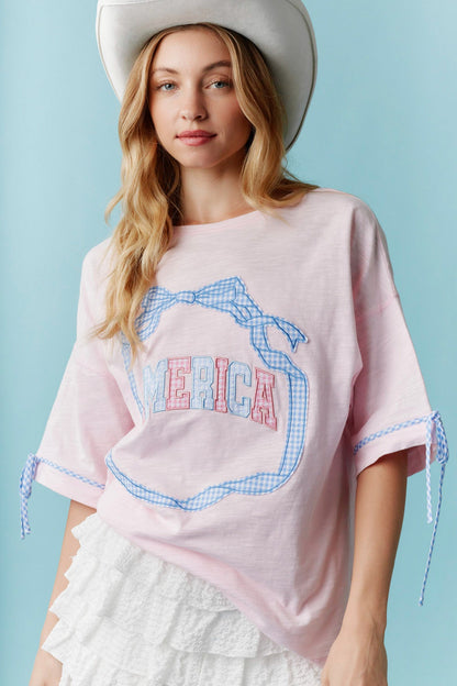 Americana Gingham Bow T-Shirt features blue gingham trim, bold “MERICA” lettering, and a bow accent. Sizes available: S, M, L, XL.