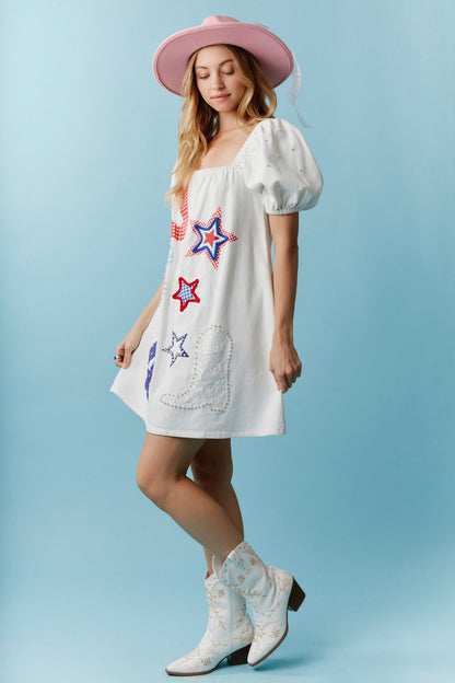 Star-Spangled Sweetheart Denim Dress with Pearl Details features puffy sleeves, colorful star patches, and embroidery. Available in sizes XS–XL.